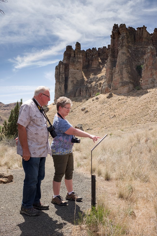 Two people read a sign along a trail. A large cliff dominates the skyline behind them.