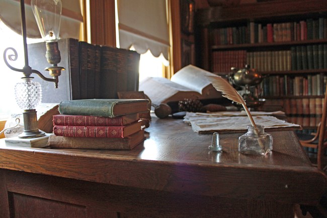 A desk by a window. Multiple items. Quill pen, books. paper.