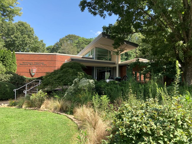 Red brick building with large tree and green garden in front. NPS Arrowhead and name of park are visible on the facade.