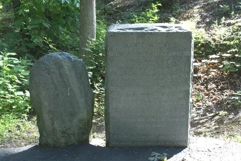 Two stone monuments are next to eachother. On the left is a crooked, smaller monument. On the right is a more modern square monument. Both have writing carved into them.