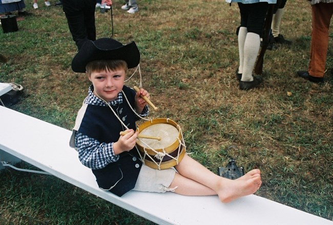 A boy sits on a concrete bench. He is holding a drum and has 18th century cloothing on.