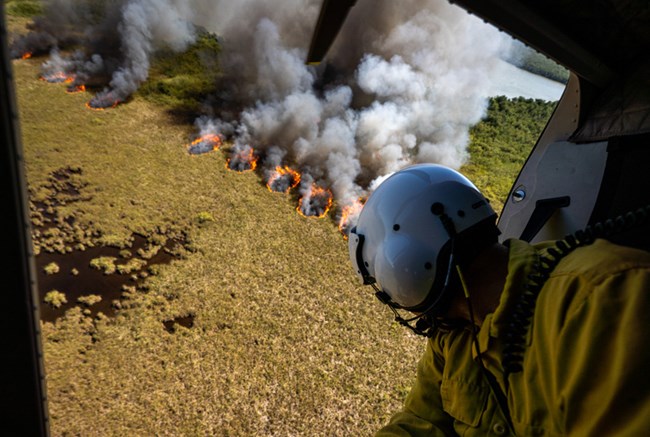 View from the inside of a helicopter with a firefighter looking out of the open door over vegetation burning on the ground.