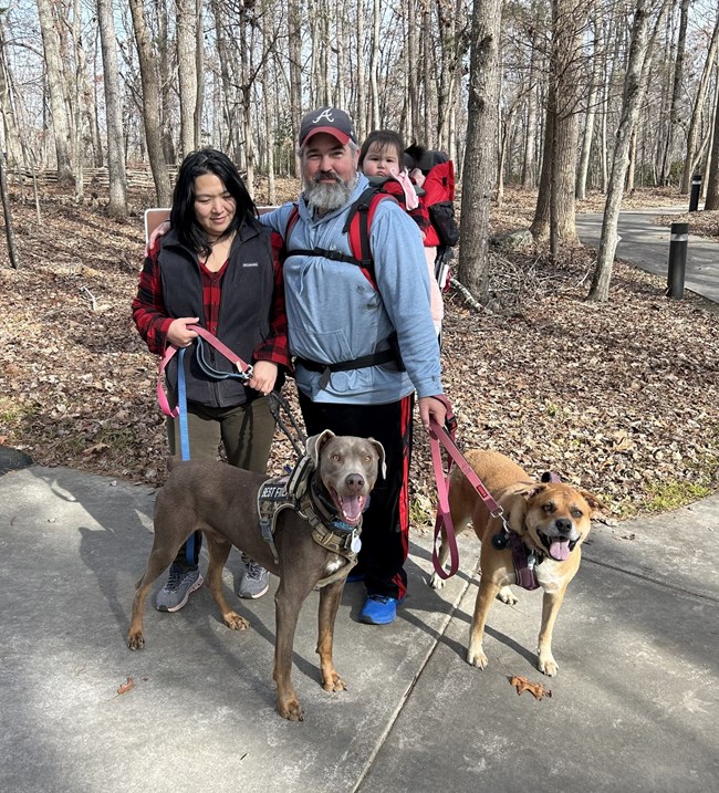 A family with two dogs is on a concrete trail surrounded by leaves and trees.