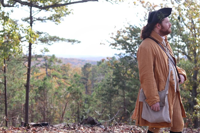 A man in 1800s clothing is looking in the distance with trees around him.