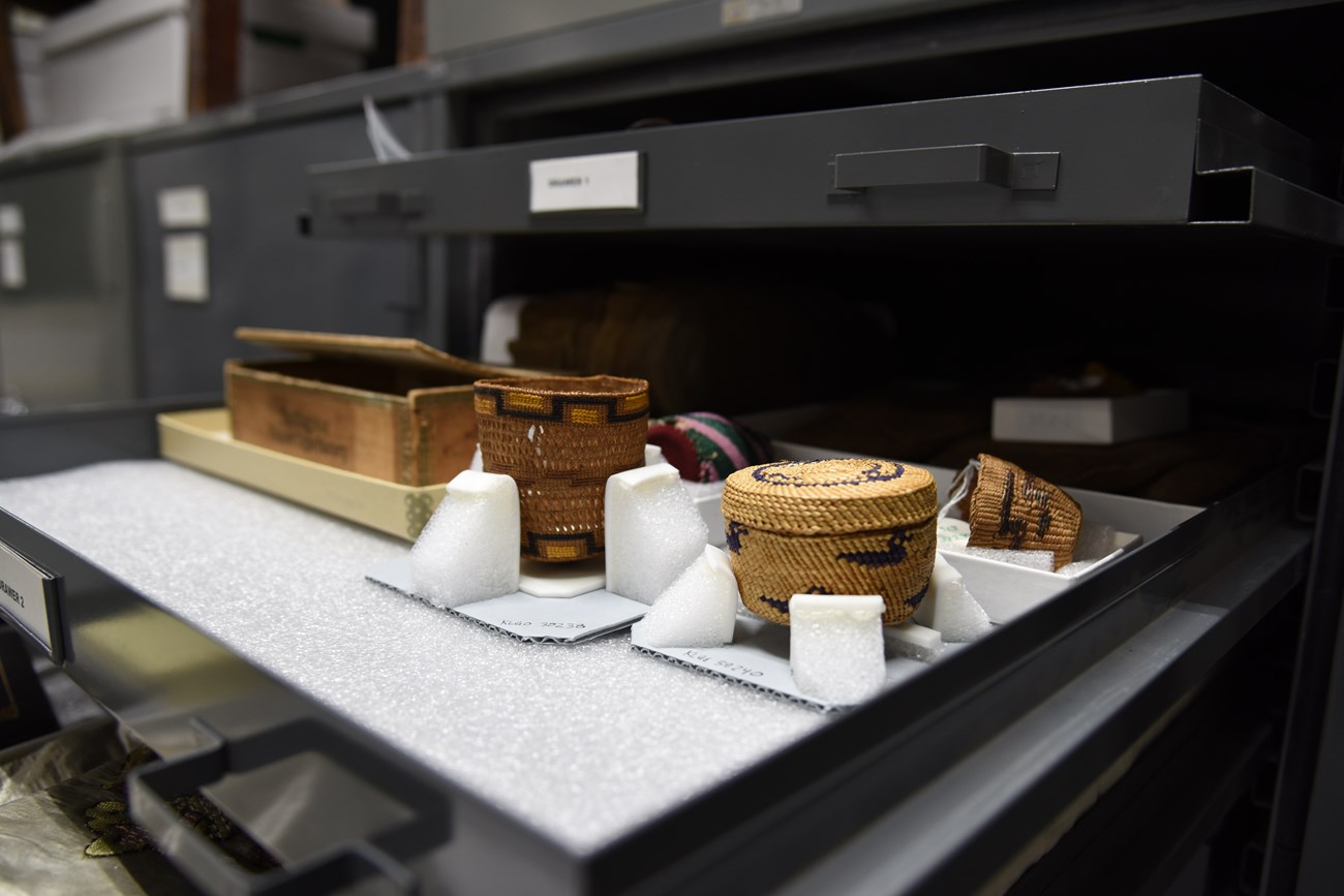 color photo of a shallow gray metal storage drawer holding open topped white boxes lined with white protective padding holding various small artifacts