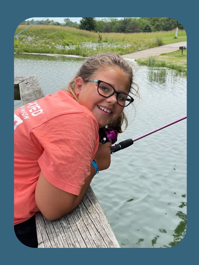 Middle schooler leaners over dock and smiles, holds a fishing pole. Pond beyond.