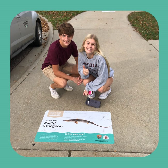 Two teenagers kneel and smile next to large sidewalk sticker. Sticker shows fish and reads "pallid Sturgeon"