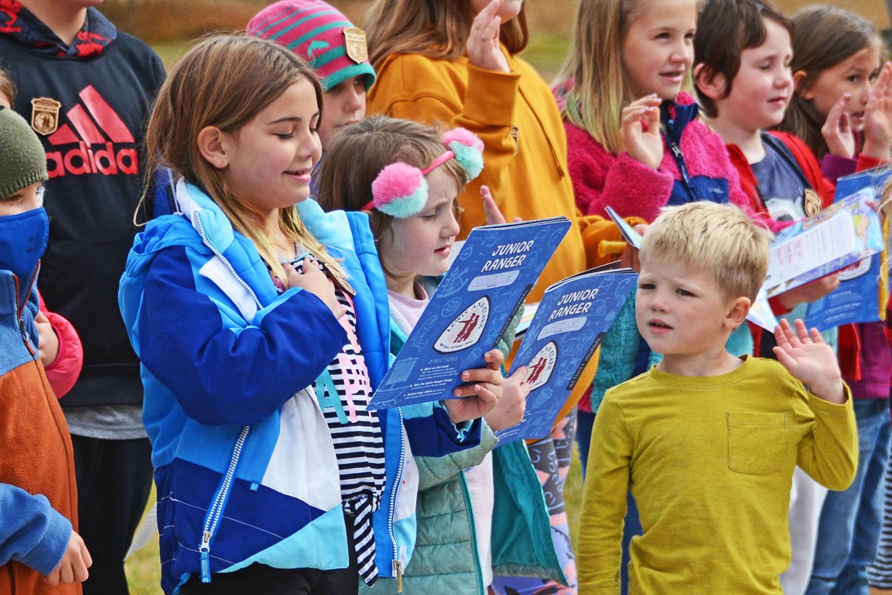 Group of children hold blue junior ranger booklets and take pledge. They smile as they read the back of the booklet. Golden trees in the background.