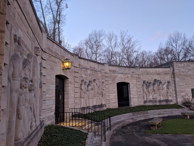 Curved limestone building with 3 sculptured panels with scenes of people, stone walkway leads to stairs and doorway, blue sky