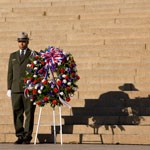 Man with green uniform and hat standing on staircase next to wreath on pedestal