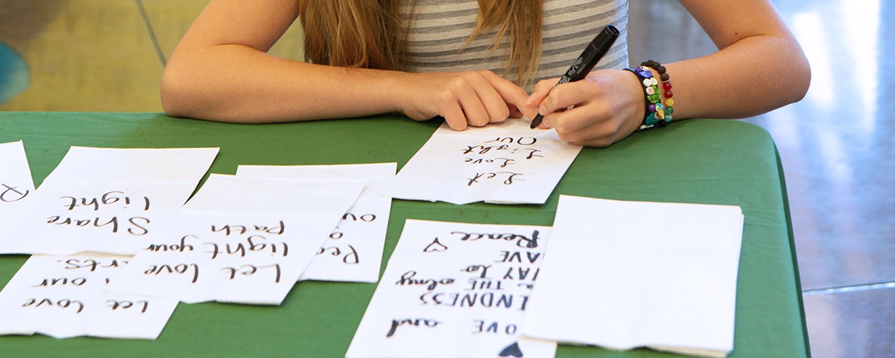 A color photo of a table with white paper bags, some with handwritten messages. Someone is writing a message on one.