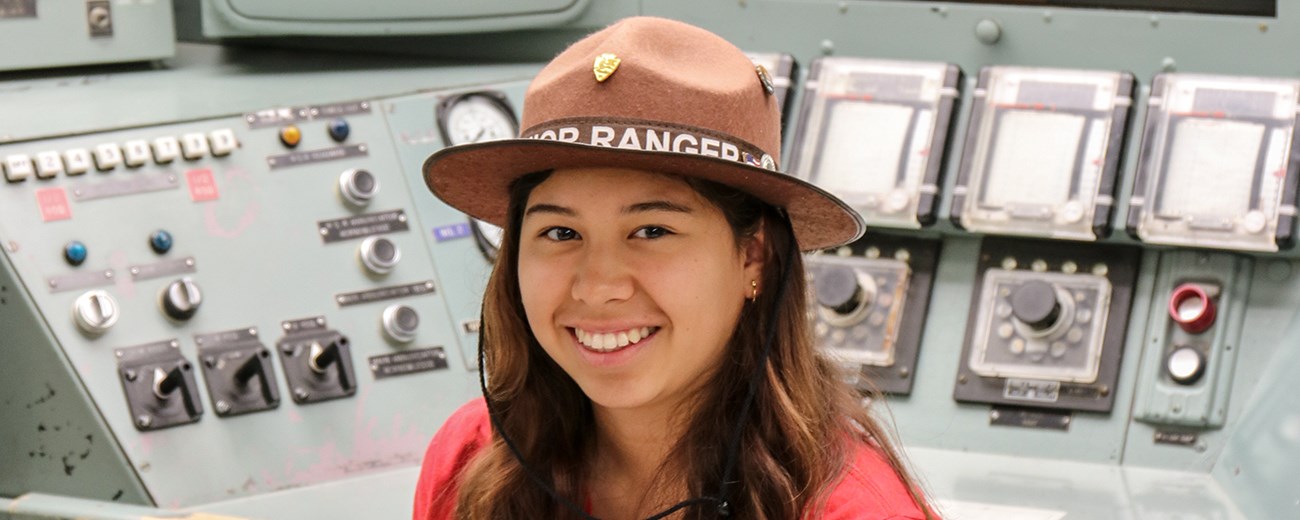 A young person sits in front of a desk with many dials and gauges.