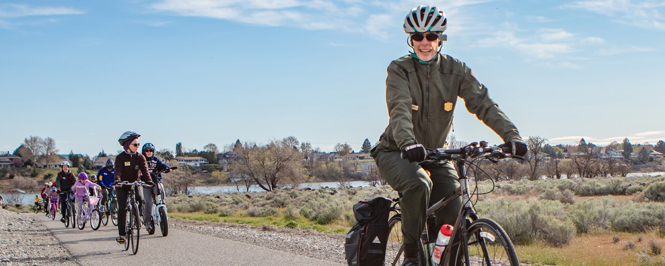 A woman in uniform on a bike wearing a helmet leads a group of adults and children riding bikes on a sunlit paved path.