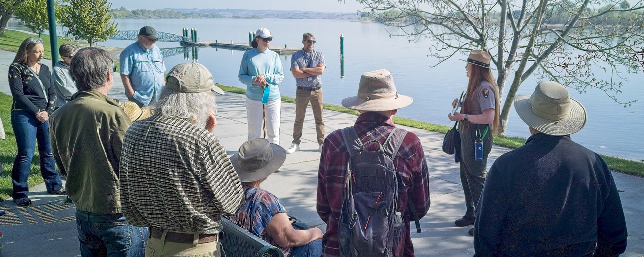 10 adults stand in a semicircle listening to a uniformed ranger along a river on a sunny day.