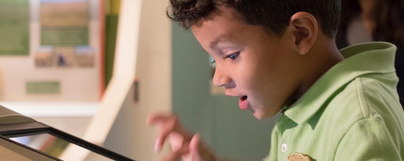 A young boy wearing a Junior Ranger badge presses on an interactive screen in a museum.