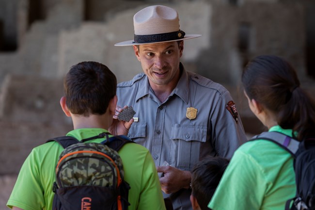 Ranger in flat hat, gray shirt with badge, green pants talks with two boys.