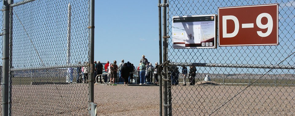 Inside a fence a ranger stands over a group of people near a metal and glass structure.