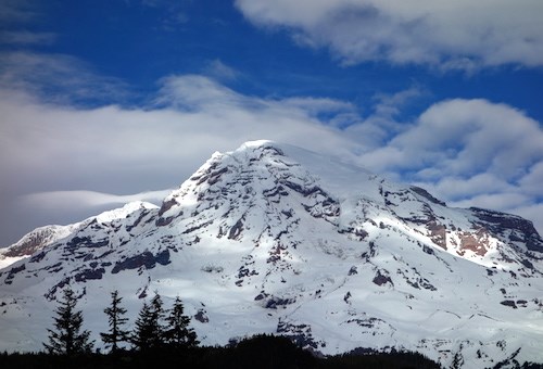 Swirling clouds cast shadows while patches of sunlight highlight ridges on a snowy mountain.