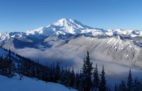 A snow covered mountain rises above a forested river valley dusted with snow.