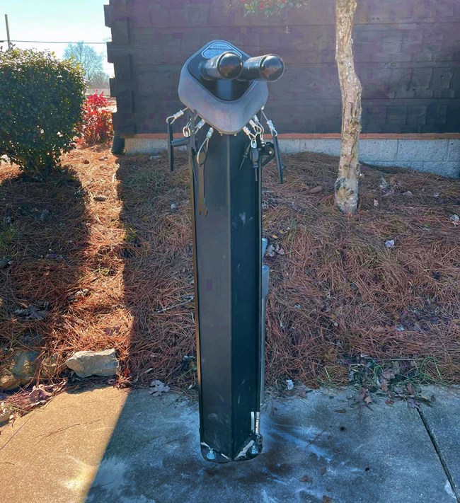 A square structure with two rods and several small tools hanging from it, at the top of a square metal pole.