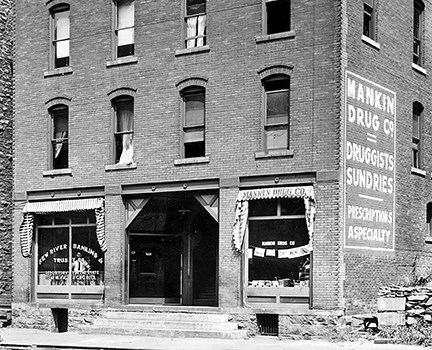 A brick building with two large glass windows and steps leading to the entrance. One window says New River Banking and Trust Company.