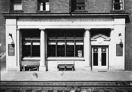 A brick building with an ornate cement facade that says National Bank of Thurmond sitting next to railroad tracks.