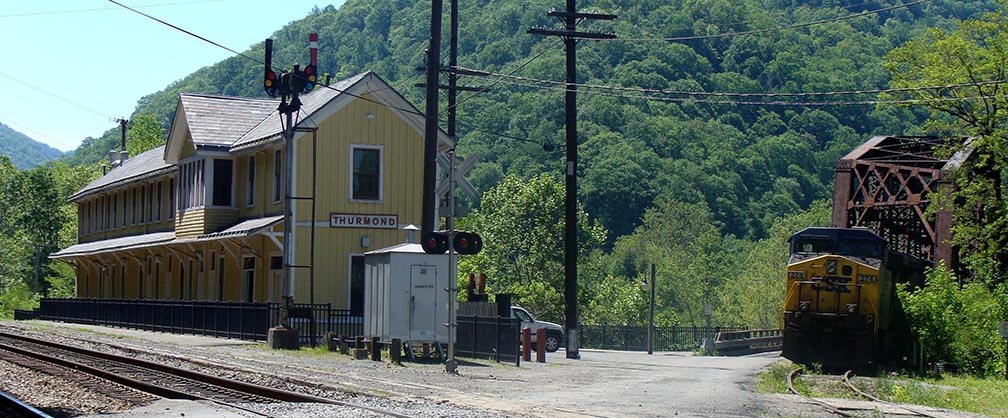 A colored photograph of the yellow Thurmond Depot Visitor Center. A train is approaching on the right side.