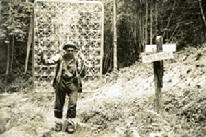 This black and white picture shows a man next to a makeshift sign that says "Humes Ranch." The man has a comically large box spring attached strung to his back.