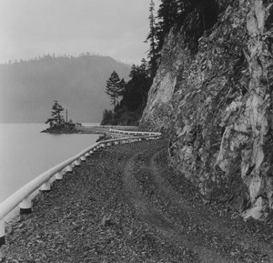 A black and white photo of a windy, dirt road wedged between a cliff to the right and a lake to the left. The road appears to be cut from the cliff. A low, olden style guard rail edges the road on the left, just above the lake.