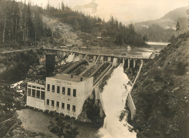 A historic photo of the Elwha Dam in 1913. Water emerges from the dam via designated channels on each side of a powerhouse structure. The powerhouse lies just below the dam and has multiple tubes that draw water from the reservoir.