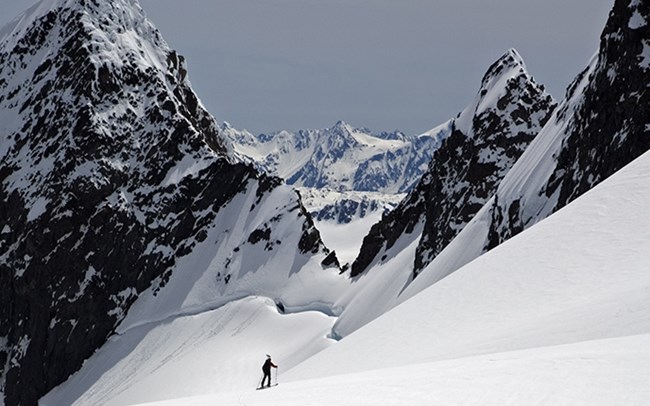 A researcher traversing a snow-covered glacier on skis. Dramatic, jagged mountain peaks loom just above the glacier. More mountains and glaciers can be seen in the distance.
