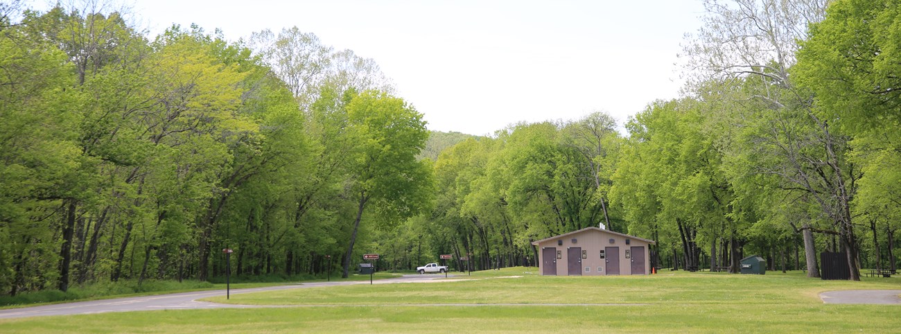 A large grass field with a showerhouse in the background. Trees line the field.