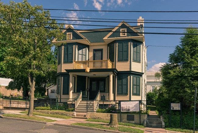 A cream-yellow two-story Victorian house with green trim & shutters - bay windows flank a porched entrance. A sign denotes it as the American Labor Museum