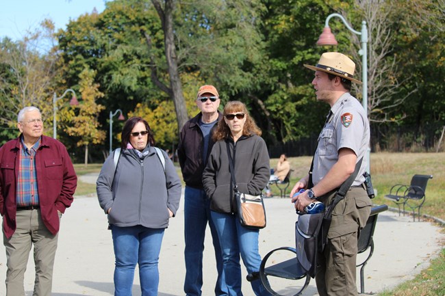 A park ranger in grey & green uniform with straw flat hat speaks with four adults