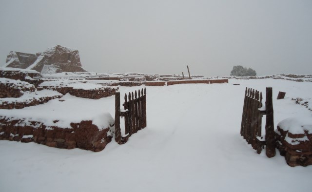 An open gate leads across a snowy space toward tall adobe structural remains with a few inches of snow on top.