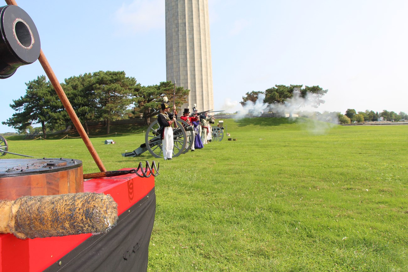 7 people in line wearing War of 1812 uniforms or period dress level muskets to fire. They are positioned around 4 different pieces of artillery. Smoke drifts down range.