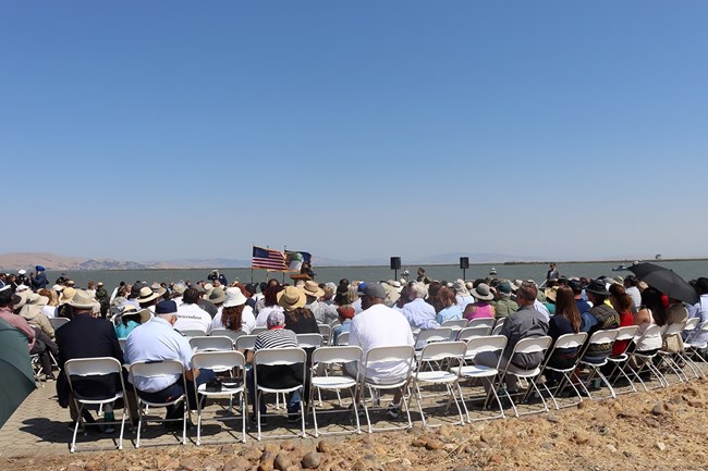 Seated crowd facing away from camera with an American flag flying behind a speaker at a podium.