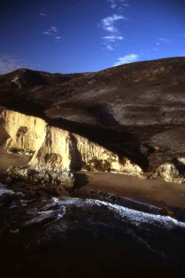 An aerial photo of burnt hills beyond steep, off-white-colored coastal bluffs.