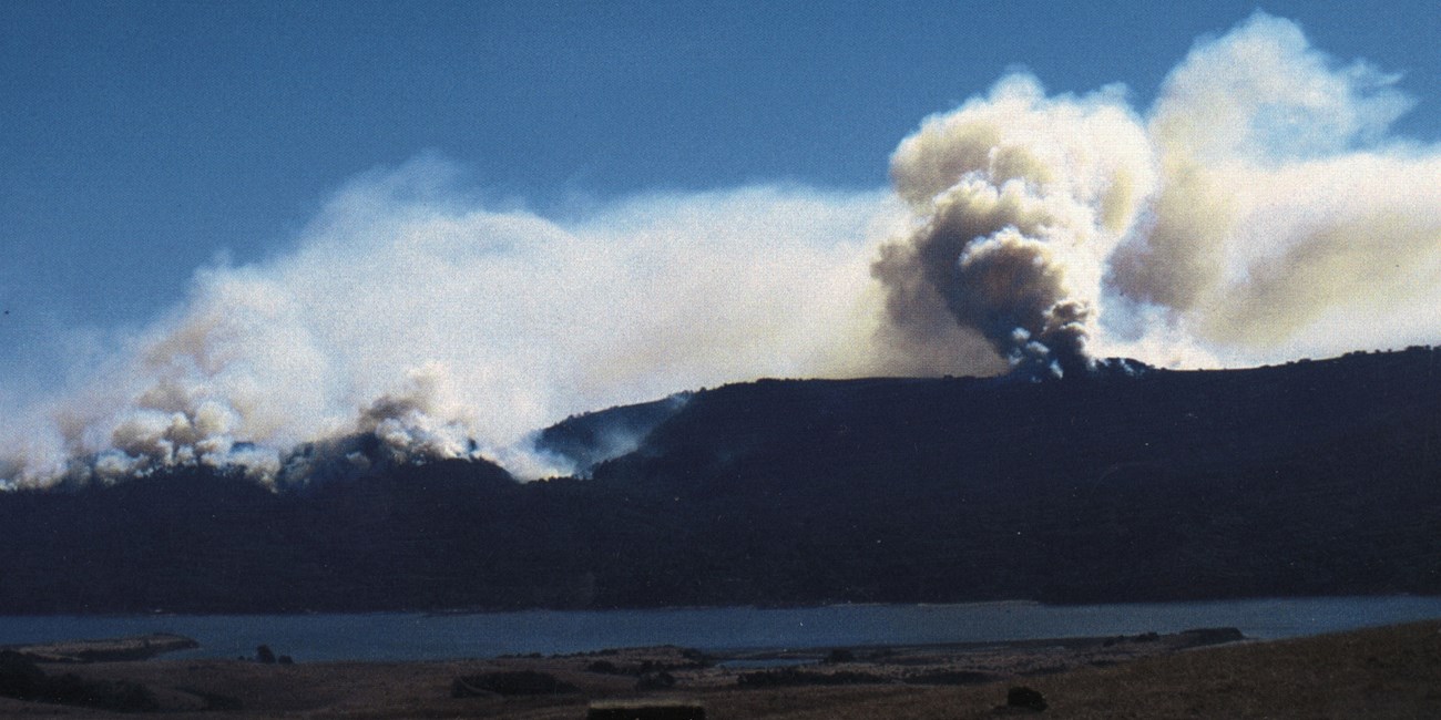 Across a narrow bay, wildfire smoke rises from a wooded ridge.