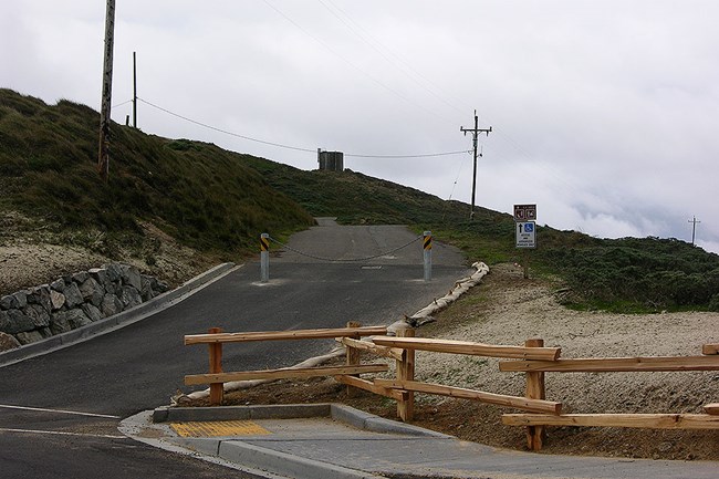 Two metal bollards connected by a heavy chain block a narrow road leading up a hill beyond a newly constructed split rail fence.