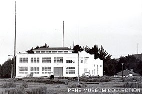 A black-and-white historic photograph of a three-story building with many windows adjacent to two radio antennae.