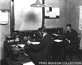 A black-and-white historic photo of two individuals sitting at desks operating Morse code equipment.