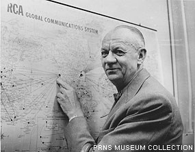 A black and white historic photograph of a man pointing to a location on a map on a wall.