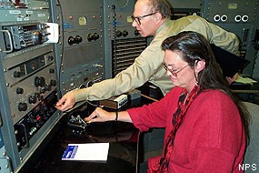 A seated woman operates radiotelegraph device as a man to her right reaches for a switch on a control panel.