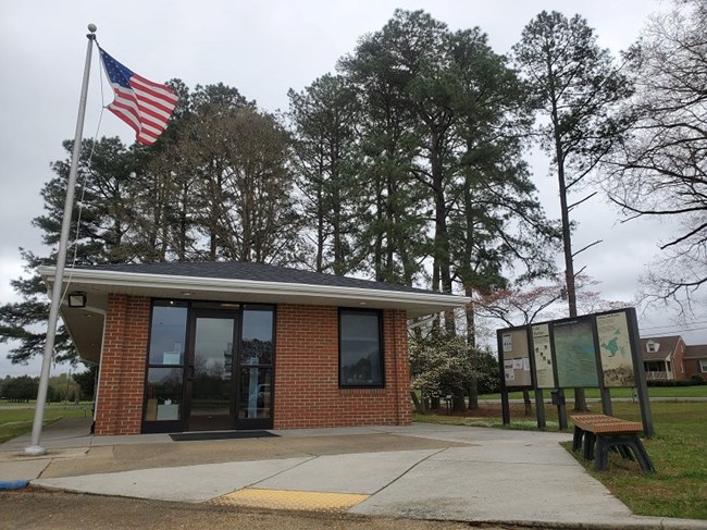 a small brick visitor center flanked by a flagpole and a freestanding interpretive sign.