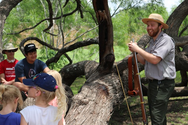 Male Park Ranger stands in front of crowd of families, holding a historic musket.