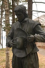 SACR_sm_Bronze-Sculture-Head-Bowed A bronze statue depicts a man in a long-sleeve cloth shirt holding a broad-rimmed hat and looking down.