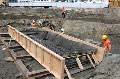 Construction workers place a wooden case around the bottom and sides of an old wooden ship.