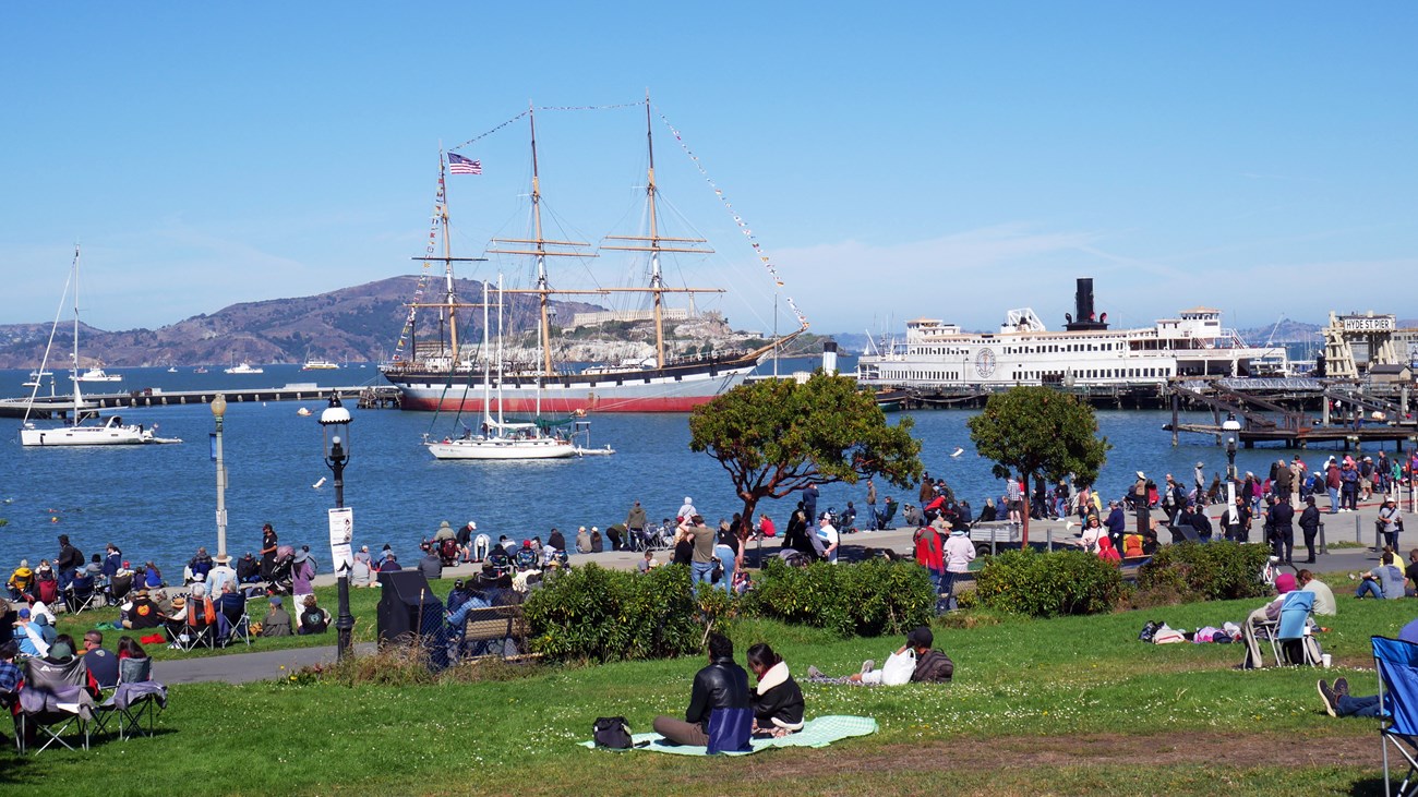 people sit on a grassy lawn and look out over Aquatic Park Cove with Balclutha in the background