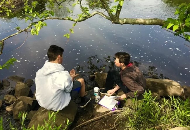 Two young people sit on a forested river bank with their backs to the camera.